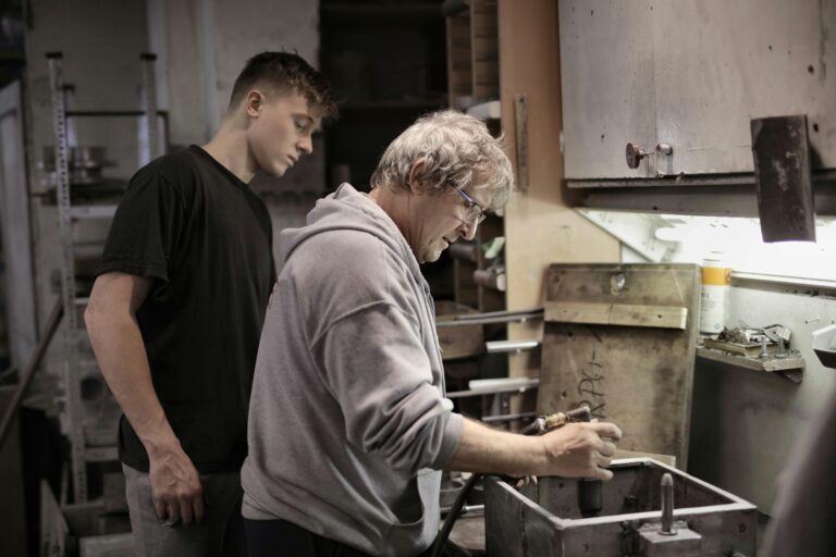 An experienced craftsman teaches a young apprentice in an industrial workshop setting.