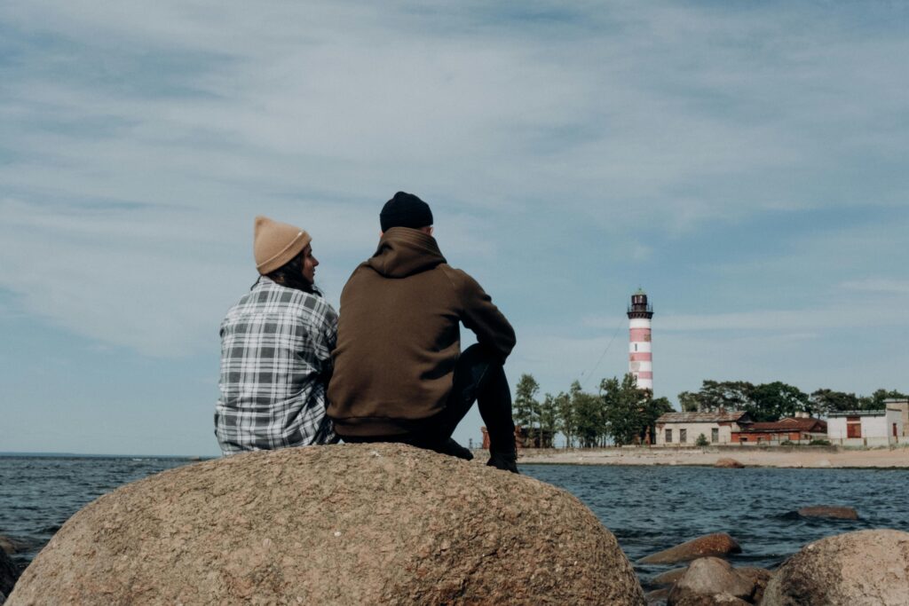 A couple sits on a rock by the sea, gazing at a lighthouse under a clear sky.